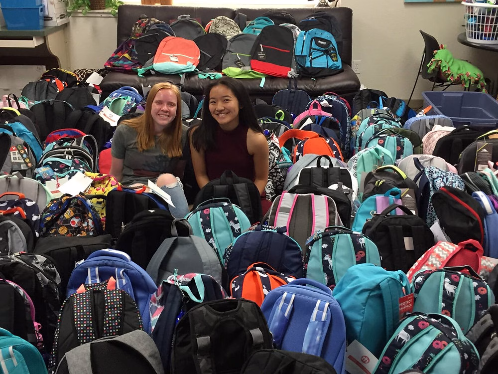 Two women sitting among a large collection of backpacks