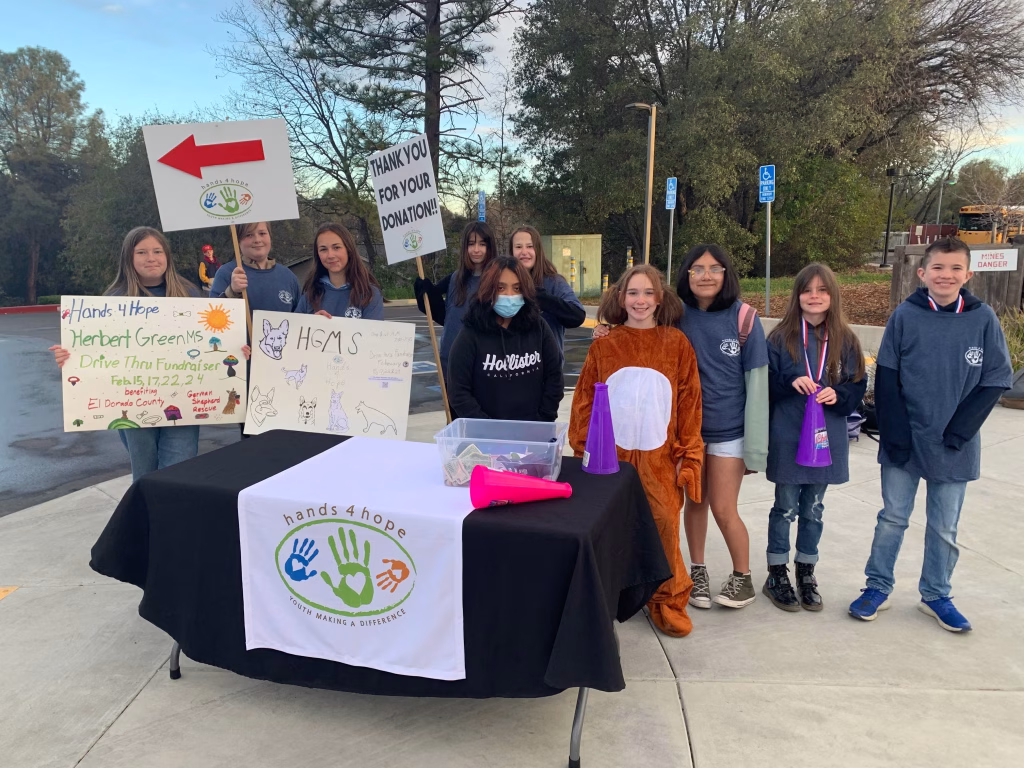 Group of children standing behind a table with signs and a tablecloth with the Hands 4 Hope logo