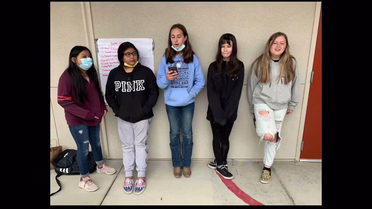 Five girls standing in a line, wearing casual clothing and masks, posing for a photo.