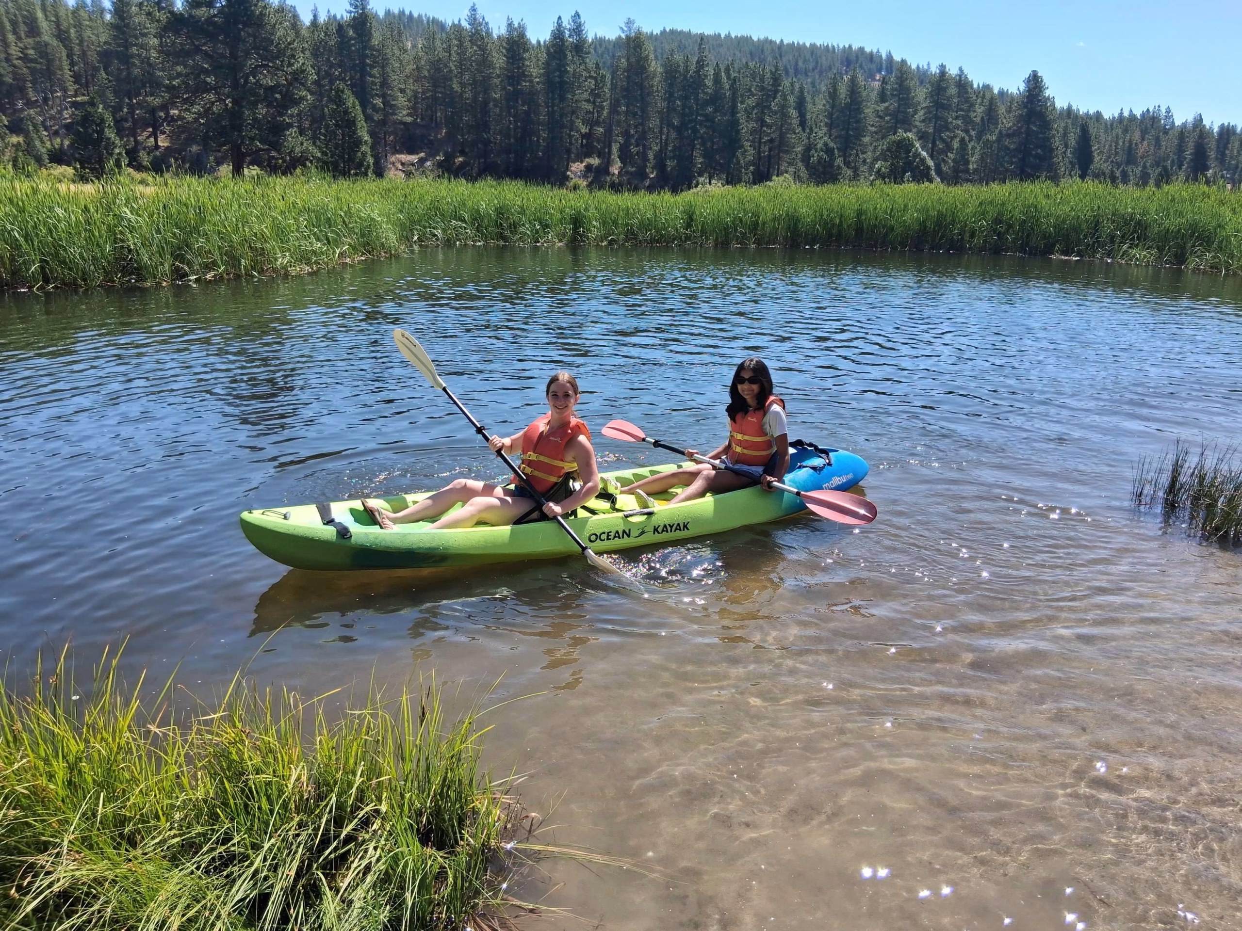 Two women kayaking in a serene lake, surrounded by lush greenery and tall trees in the background.
