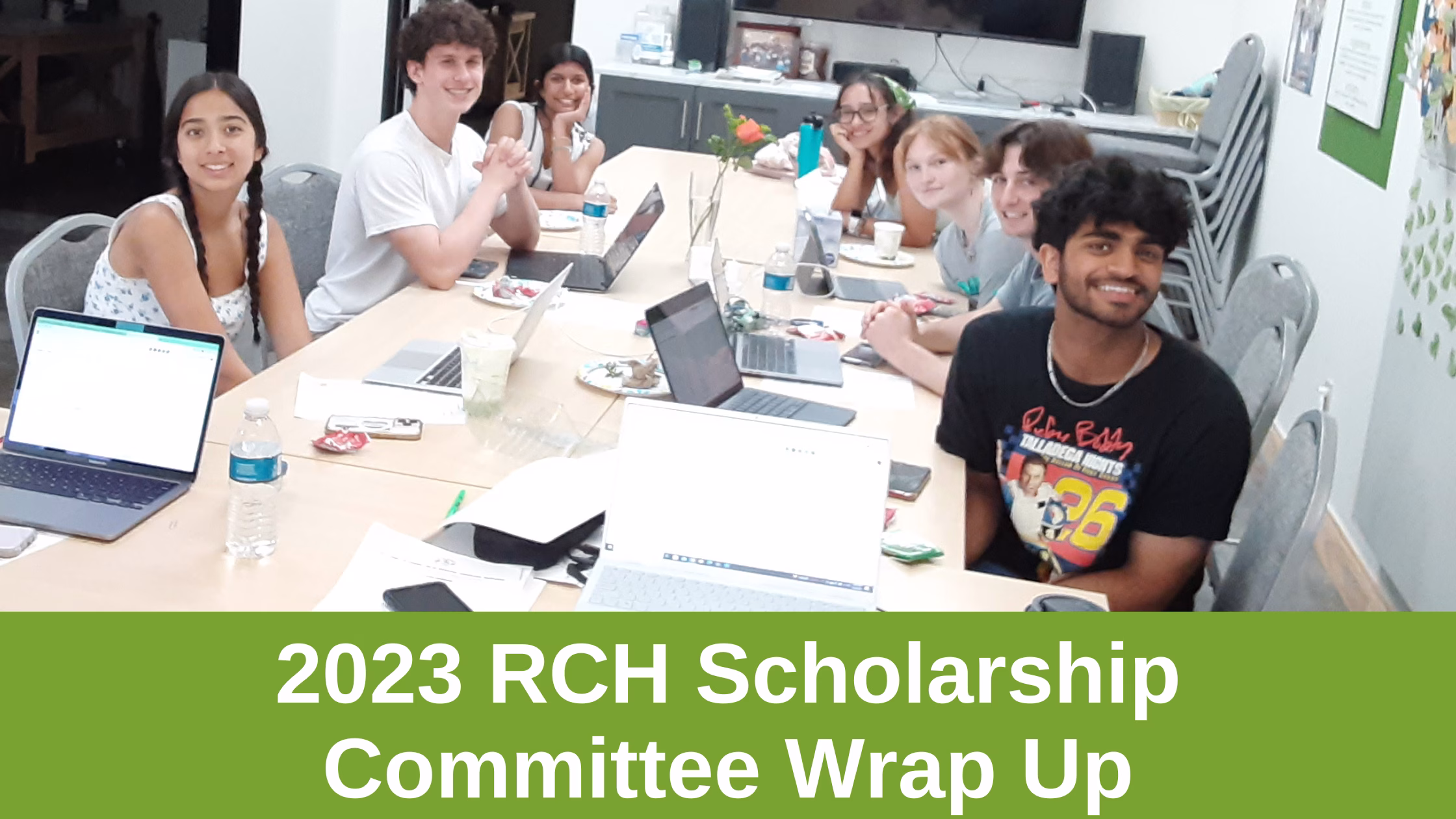 A group of students sitting around a table with laptops and papers, smiling at the camera.