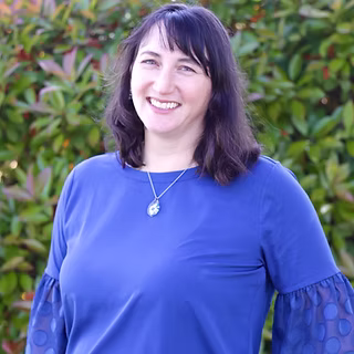 A woman with dark hair and a blue top stands in front of a backdrop of green leaves, exuding a sense of serenity and connection with nature.