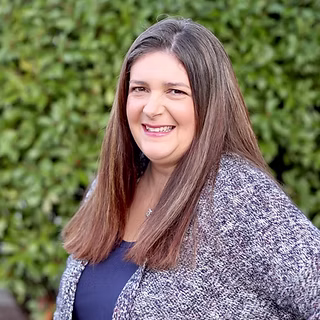 A woman with long brown hair and a gray cardigan standing in front of a green hedge.