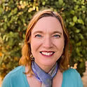 A woman with red hair and a blue scarf standing in front of a green hedge.